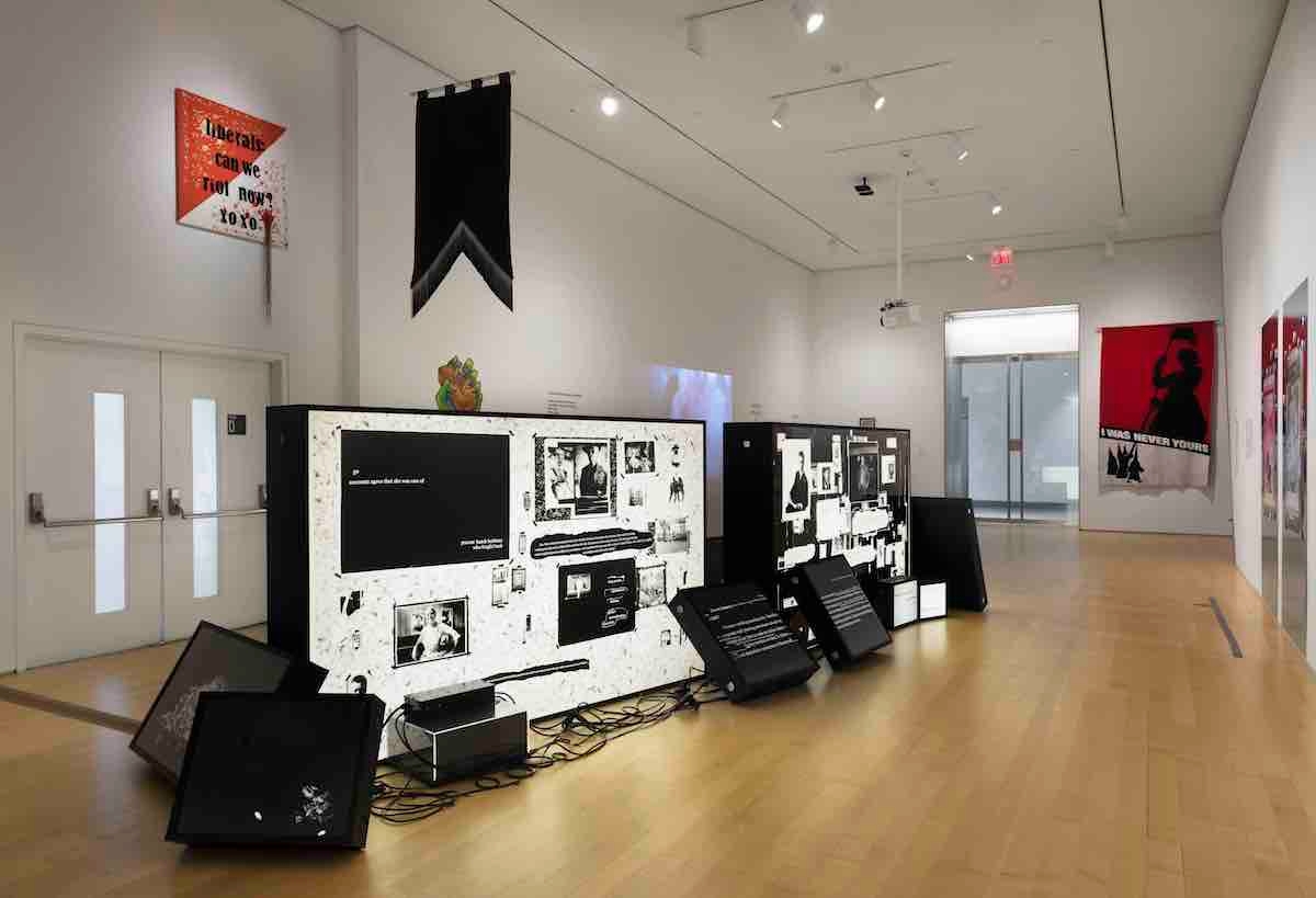 a view of the exhibition in which back light panels are covered with black and white items, a banner hangs from a wall and several red and white visual artworks hang on walls
